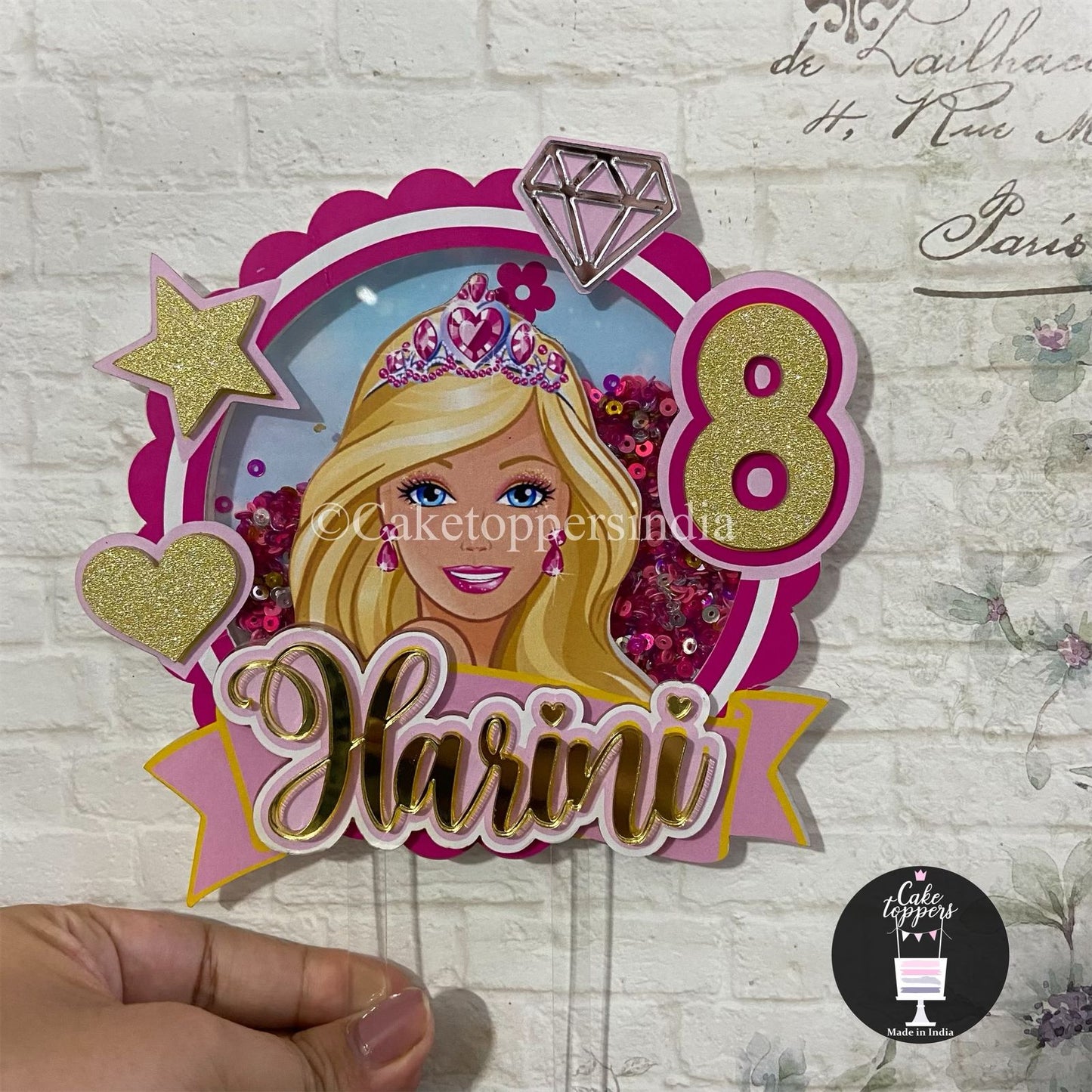 Customized Barbie theme cake topper with the customised name, featuring Barbie in a pink outfit with a crown, surrounded by stars and hearts, holding a diamond-shaped item, with the number and the name displayed in a banner.
