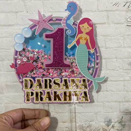 A customized cake topper featuring a mermaid theme with  customised name , including decorations such as stars and bubbles, in pink, purple, and green colors.